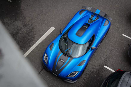 An overhead view of a striking blue Koenigsegg Agera R, showcasing its aerodynamic design and sleek features against a backdrop of parked vehicles.