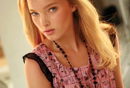 Close-up of Swedish model Elsa Hosk with blue eyes and blonde hair, wearing a pink patterned top and black beaded necklace in a softly lit setting.