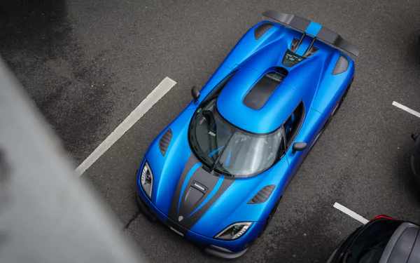 An overhead view of a striking blue Koenigsegg Agera R, showcasing its aerodynamic design and sleek features against a backdrop of parked vehicles.