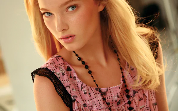 Close-up of Swedish model Elsa Hosk with blue eyes and blonde hair, wearing a pink patterned top and black beaded necklace in a softly lit setting.