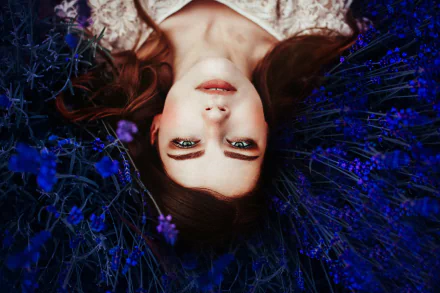 Portrait of a brunette woman with green eyes lying among blue flowers, captured in a moody 4K Ultra HD desktop wallpaper.