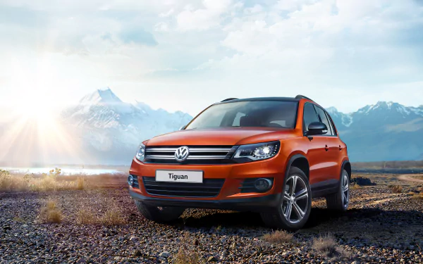Orange Volkswagen Tiguan SUV parked on rocky terrain with mountains and a bright sky in the background, captured as a HD PC desktop wallpaper and background.