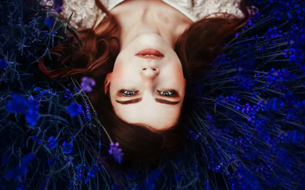 Portrait of a brunette woman with green eyes lying among blue flowers, captured in a moody 4K Ultra HD desktop wallpaper.