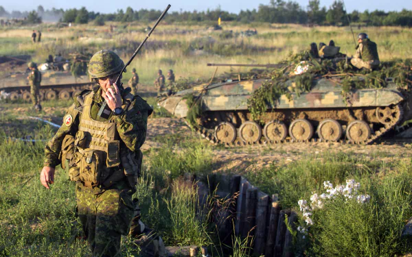A soldier communicating on a walkie-talkie stands in the foreground, with military tanks and personnel in the background, set in a grassy landscape. HD desktop wallpaper image.