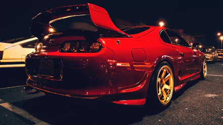 A vibrant red Toyota Supra parked at night, showcasing its sleek design and gold wheels, serves as an eye-catching HD desktop wallpaper and background.