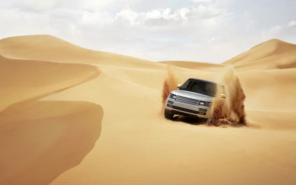 Silver Range Rover SUV drives aggressively through golden sand dunes in a desert, kicking up dust under a partly cloudy sky in this HD desktop wallpaper.