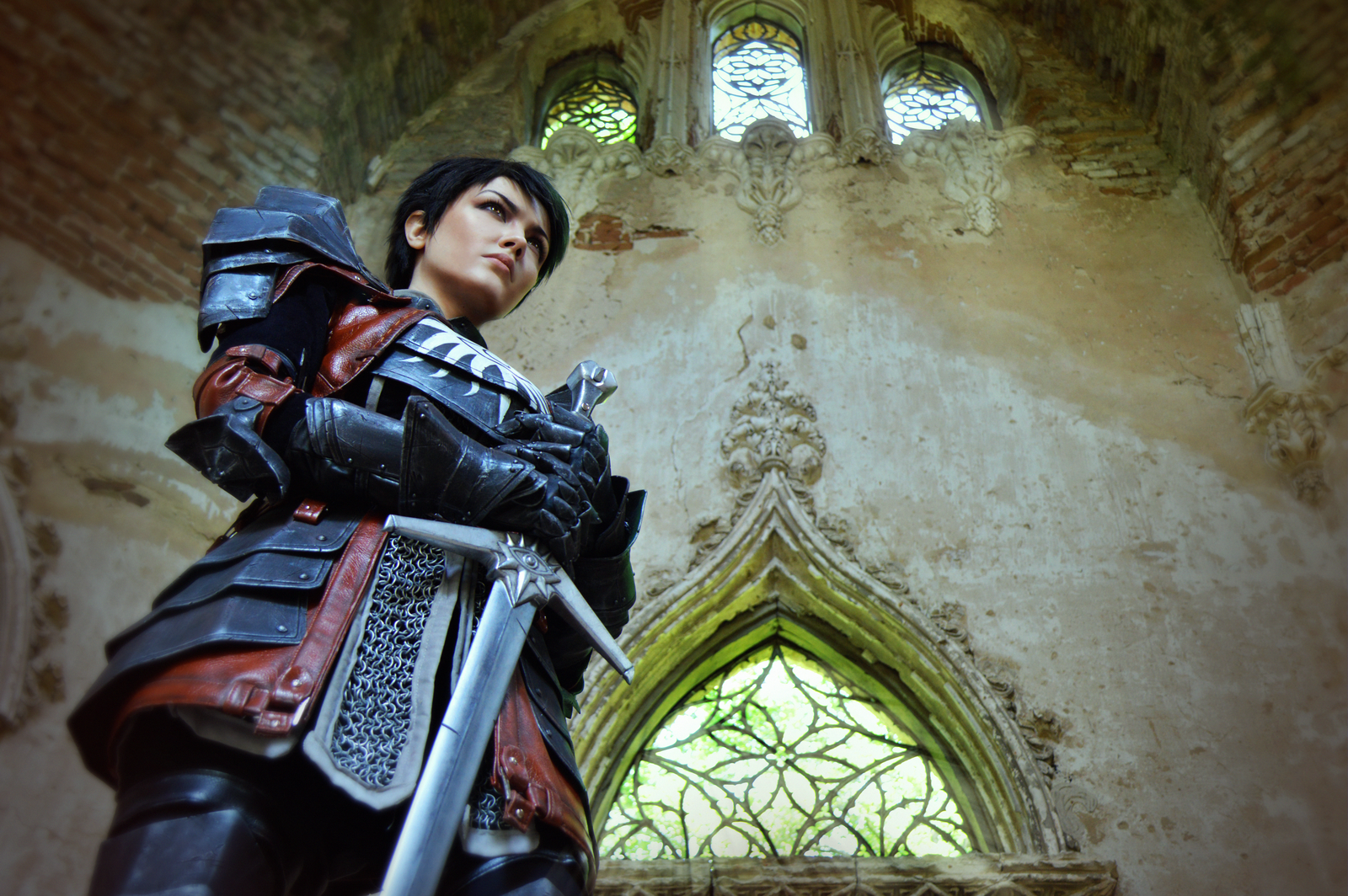 HD PC desktop wallpaper featuring a brunette woman cosplaying Cassandra Pentaghast from Dragon Age, standing in armor inside a stone building with stained glass windows.