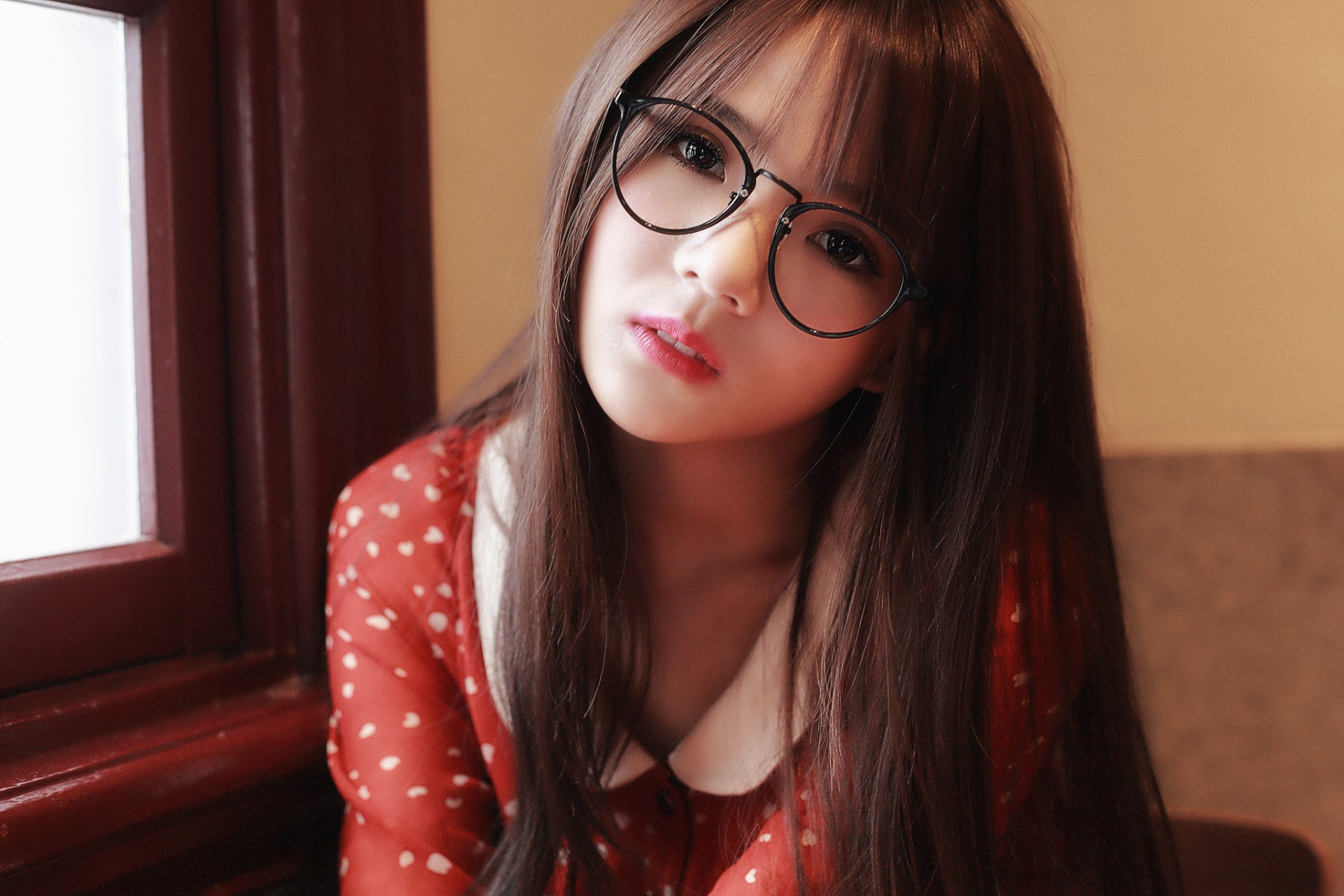 A high-definition desktop wallpaper featuring an Asian woman with brunette hair, brown eyes, and glasses, dressed in a red polka dot dress and posing indoors.