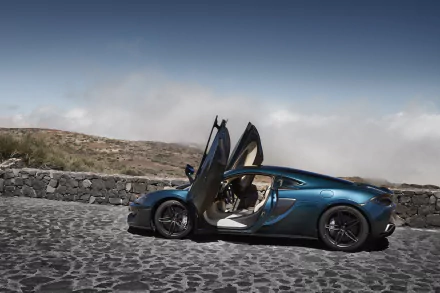 A blue McLaren 570GT with its butterfly doors open, parked on a cobblestone road against a mountainous backdrop, presented as a 4K Ultra HD PC wallpaper.