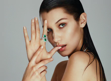 Close-up of American model Bella Hadid with blue eyes and brunette hair, posing with an emerald ring against a neutral background in an HD desktop wallpaper style.