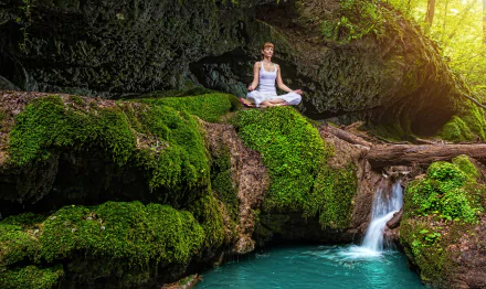 A serene HD desktop wallpaper featuring a woman practicing yoga on a moss-covered rock above a tranquil pond and small waterfall.