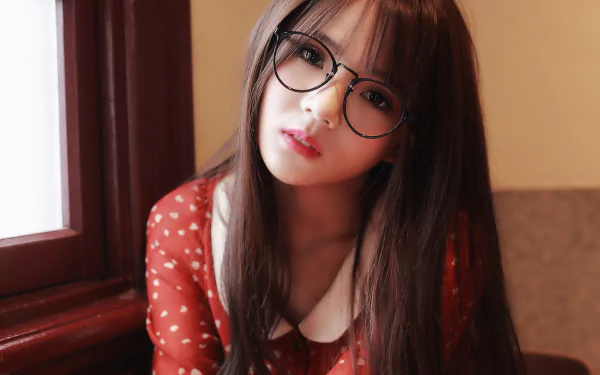 A high-definition desktop wallpaper featuring an Asian woman with brunette hair, brown eyes, and glasses, dressed in a red polka dot dress and posing indoors.