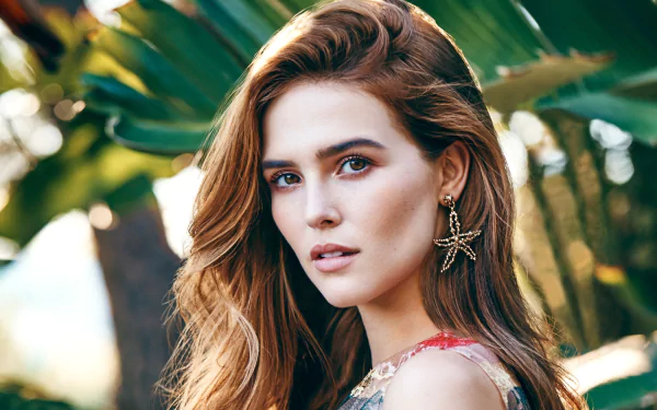 Close-up portrait of American actress Zoey Deutch with brunette hair, brown eyes, and starfish-shaped earrings, set against a lush green background.