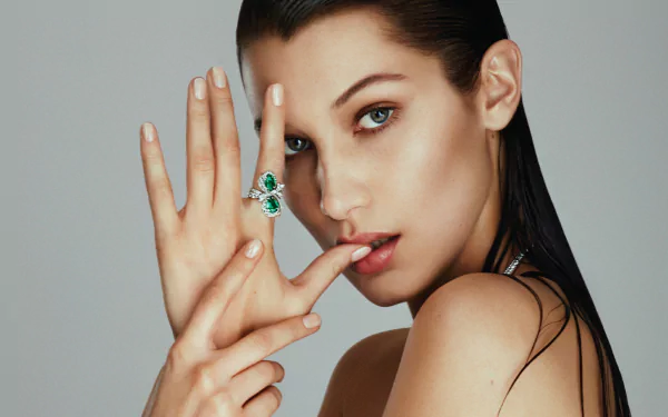 Close-up of American model Bella Hadid with blue eyes and brunette hair, posing with an emerald ring against a neutral background in an HD desktop wallpaper style.