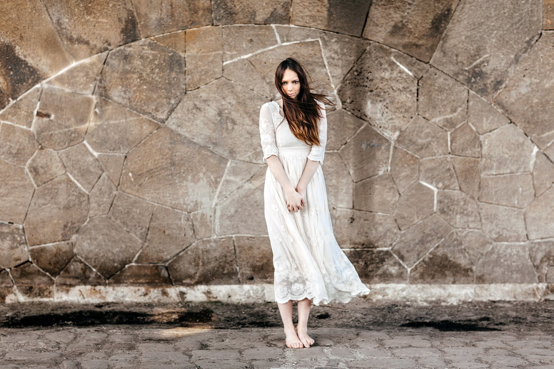 Brunette woman in a flowing white dress stands barefoot against a textured stone wall, captured in a high-definition PC desktop wallpaper and background.