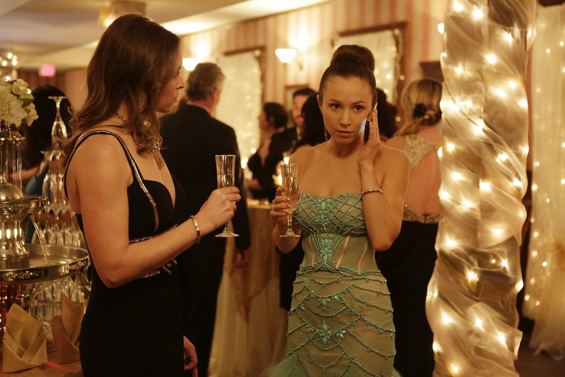 Wynonna Earp TV show scene: two women in formal dresses at a twinkling party; central woman in a green gown holds champagne and a phone — 2K Quad HD PC desktop wallpaper.