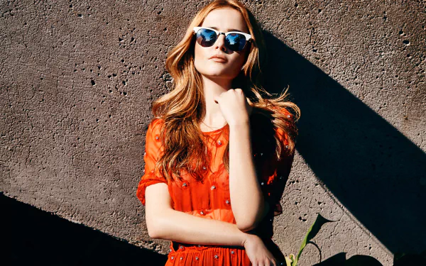 Brunette woman in sunglasses and a red dress posing against a sunlit textured wall — crisp HD desktop wallpaper.