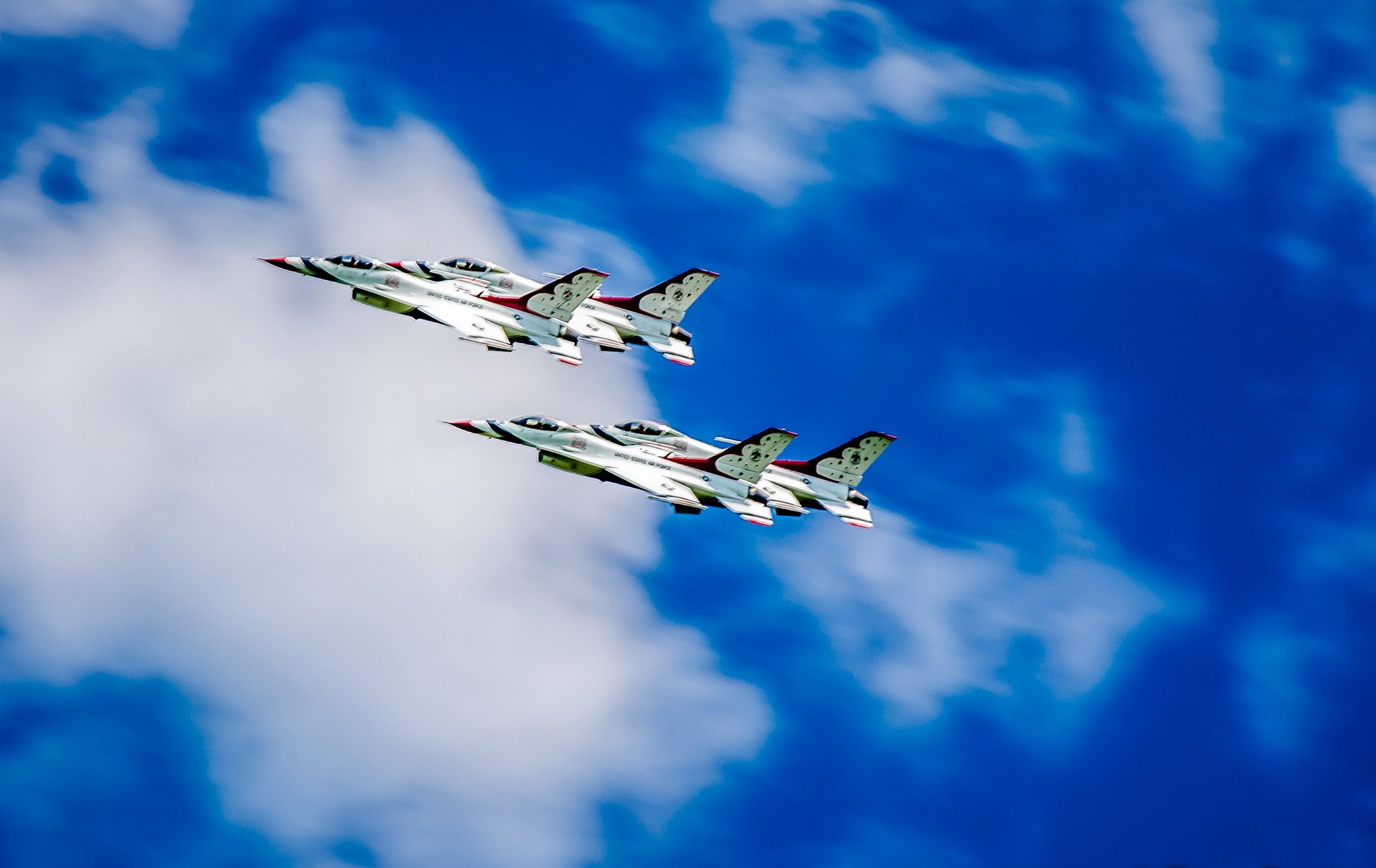 Two United States Air Force Thunderbirds jets perform at an air show, captured with motion blur against a blue sky with scattered clouds.