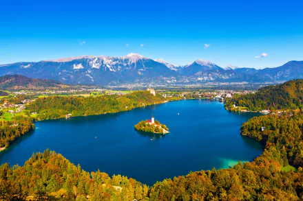 Aerial view of Lake Bled in Slovenia featuring the Assumption of Mary Church on a small island, surrounded by mountains and lush green landscape under a clear blue sky.