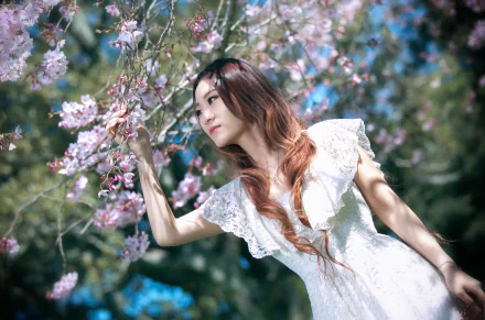 A brunette woman with brown eyes wears a white dress, gently touching cherry blossoms in a vibrant outdoor setting, captured in stunning 4K Ultra HD detail.