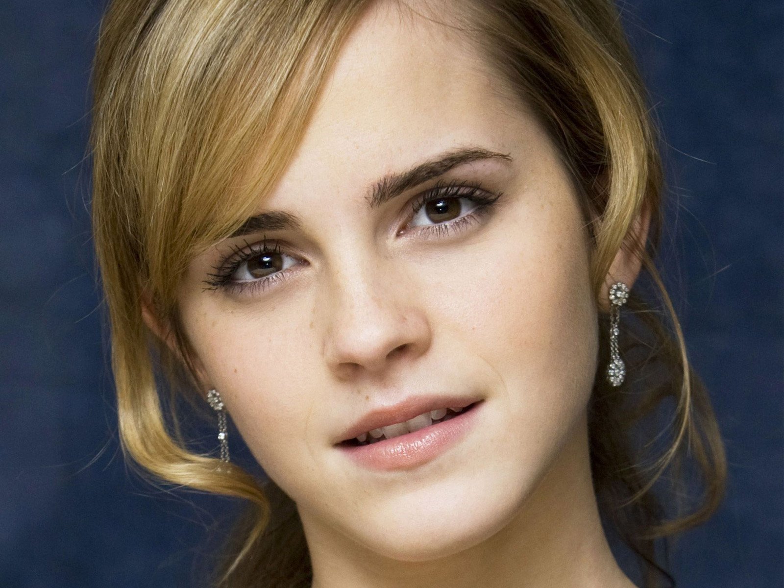 Close-up HD desktop wallpaper of celebrity Emma Watson with soft makeup and elegant earrings against a blurred dark blue background.
