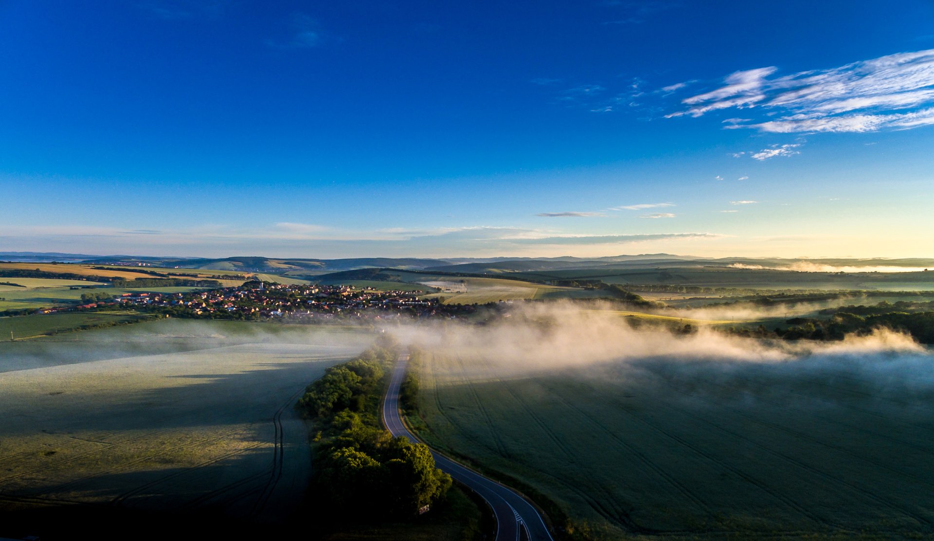 Panoramic HD landscape photography of a Czech Republic village at horizon with winding road, early morning fog, and a clear blue sky, captured as a desktop wallpaper.