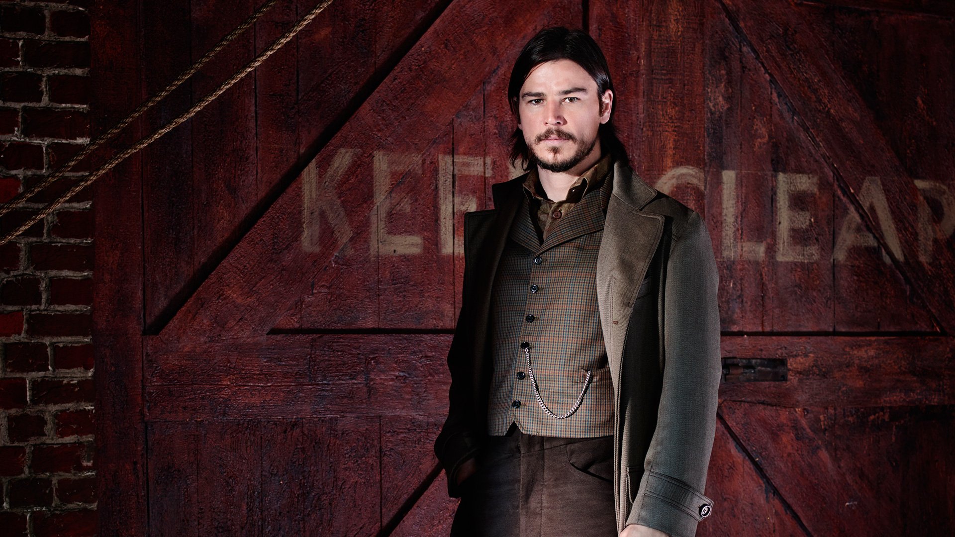 A character from the TV show Penny Dreadful stands confidently in front of a rustic red backdrop, exuding a sense of mystery and charm. High-definition desktop wallpaper for fans.