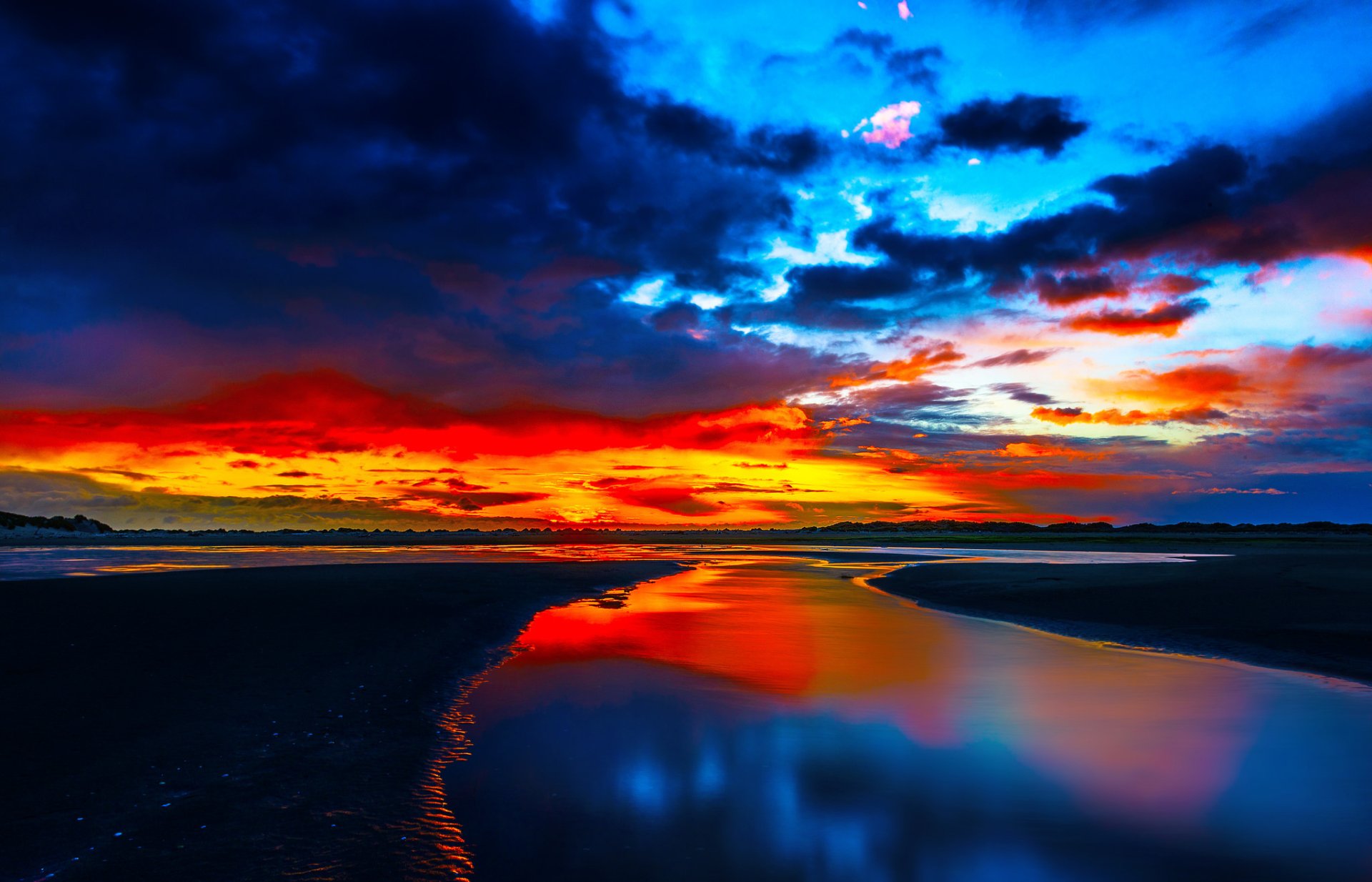 HD PC desktop wallpaper: vivid sunset sky with dramatic clouds over a coastal landscape at dusk, glowing horizon and night-hued colors mirrored in calm water reflection, a nature background.
