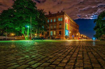 A vibrant night scene in Berlin, showcasing a man-made building illuminated by colorful lights, with cobblestone streets and lush trees lining the atmospheric city street.