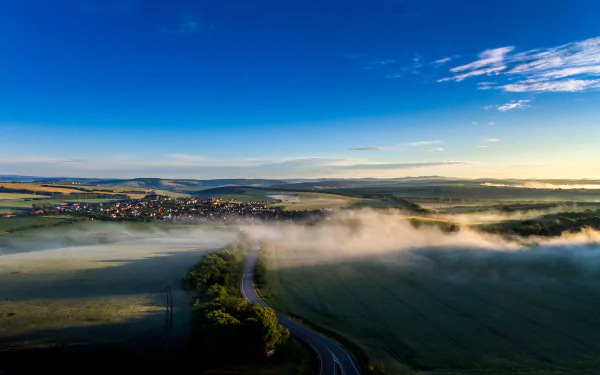 Panoramic HD landscape photography of a Czech Republic village at horizon with winding road, early morning fog, and a clear blue sky, captured as a desktop wallpaper.