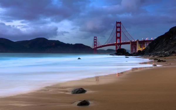 A serene beach scene featuring the iconic Golden Gate Bridge, with gentle waves lapping at the sandy shore and a dramatic sky enhancing this stunning HD wallpaper.
