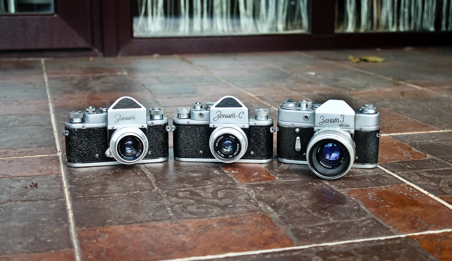 Three vintage film cameras lined up on worn terracotta tiles, a high-detail 4K Ultra HD PC desktop wallpaper highlighting textured floor and chrome camera bodies.