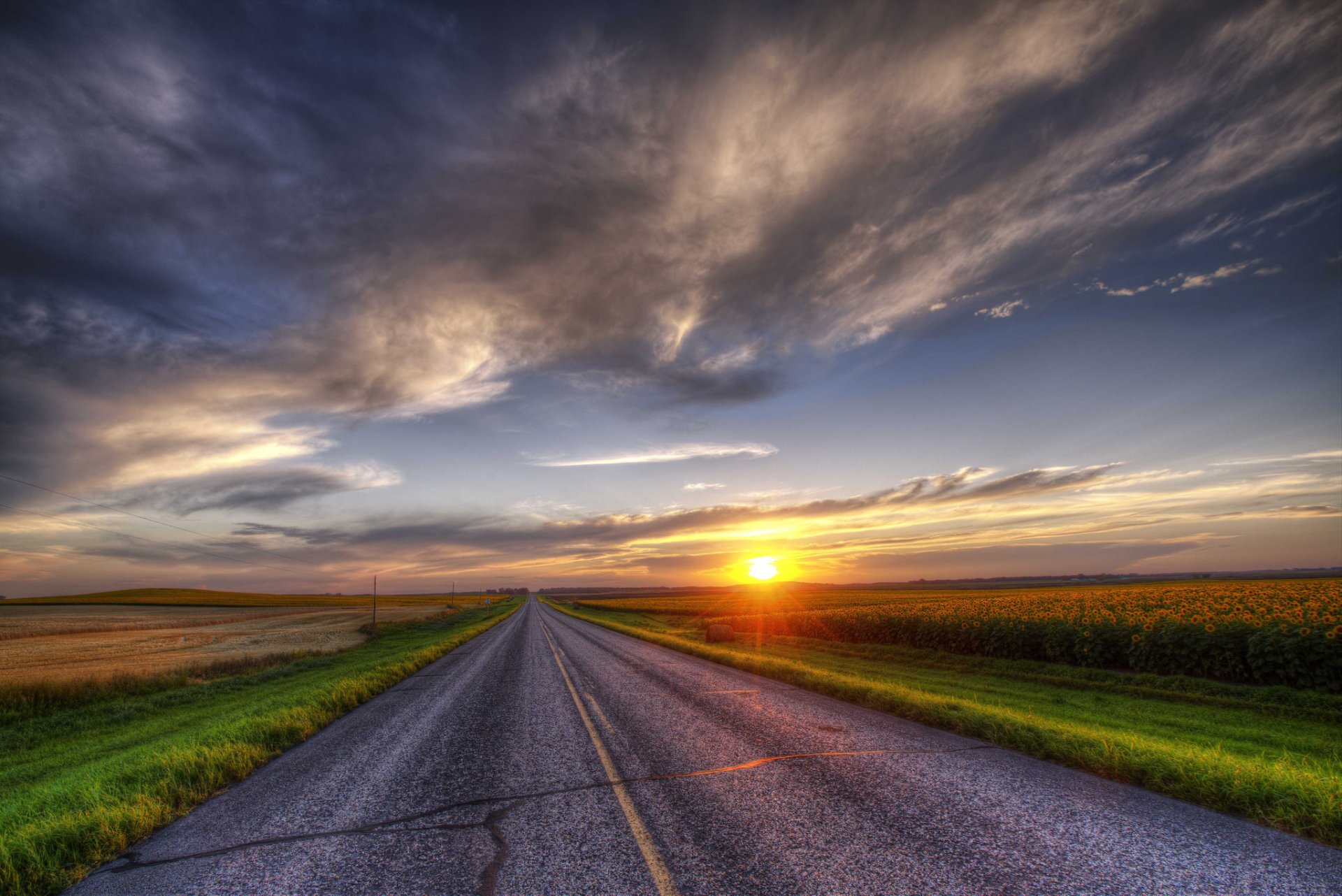 HDR HD PC desktop wallpaper - a straight man-made road through sunflower fields toward a summer sunset on the horizon, sun and dramatic sky over a wide landscape background.