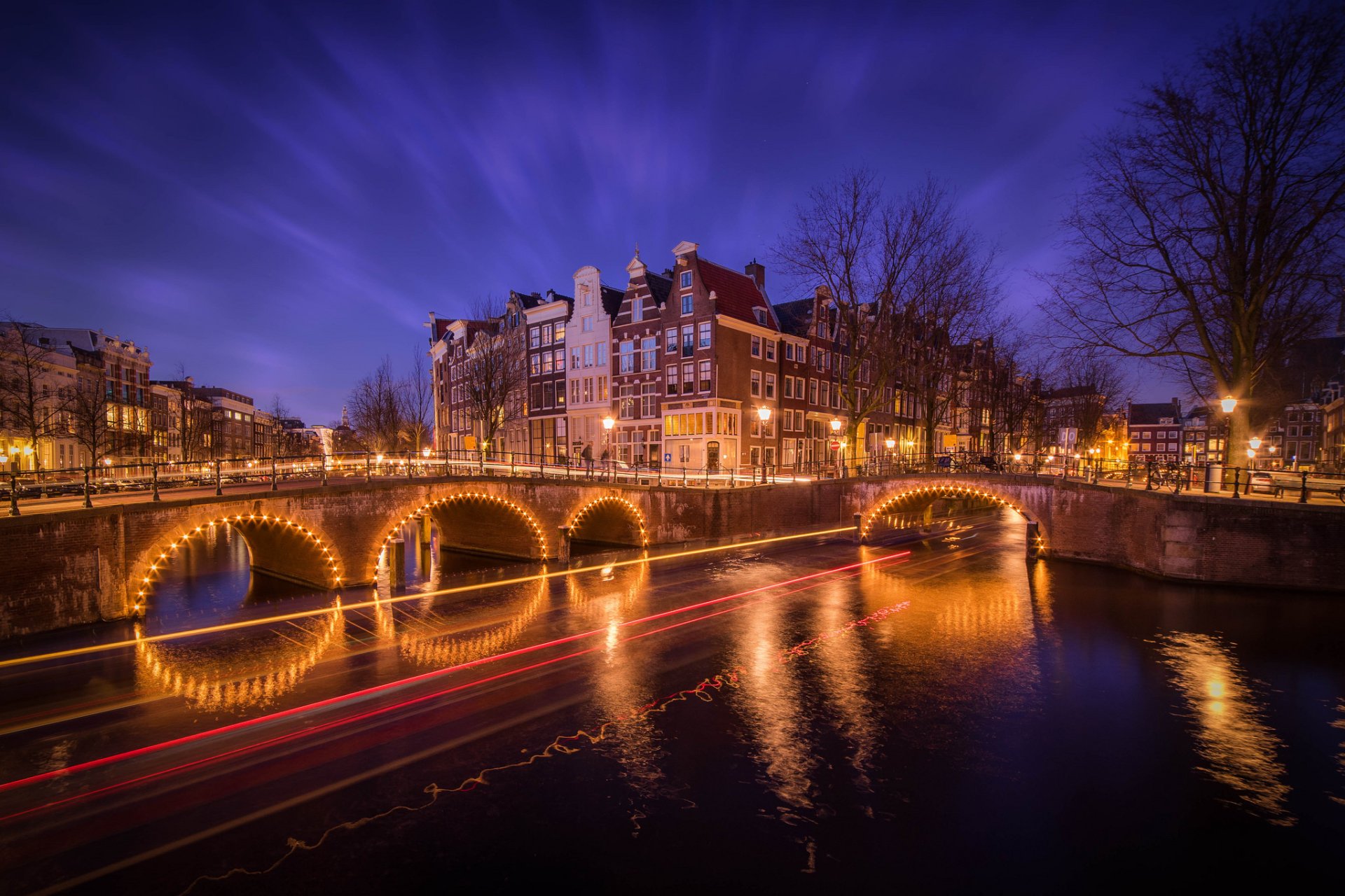 Nighttime view of Amsterdam's illuminated bridges and historic buildings reflected in the water, captured with a time-lapse effect showcasing vibrant city lights and movement.
