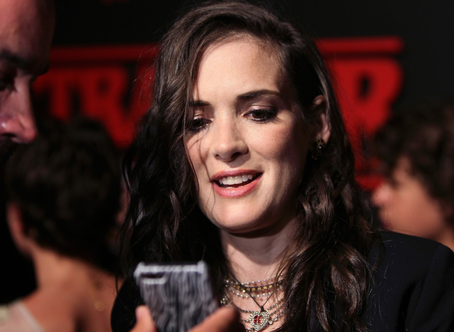 HD PC desktop wallpaper featuring a close-up of celebrity Winona Ryder smiling at a phone, with blurred people and red background lighting behind her.