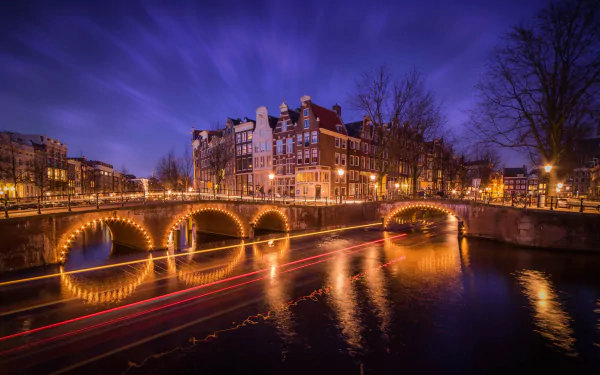 Nighttime view of Amsterdam's illuminated bridges and historic buildings reflected in the water, captured with a time-lapse effect showcasing vibrant city lights and movement.