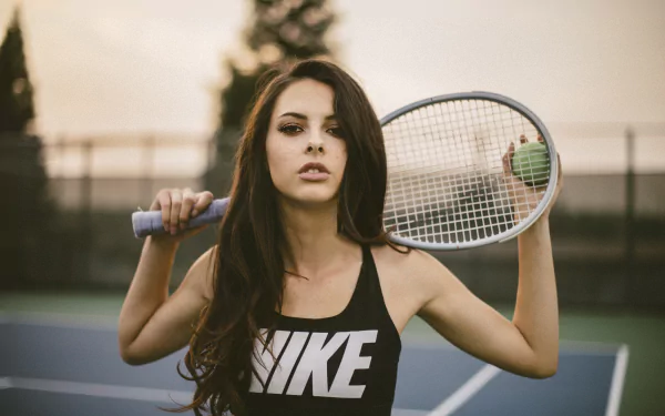 Brunette woman model with brown eyes poses confidently on a tennis court, holding a racket behind her shoulders in a 4K Ultra HD desktop wallpaper.