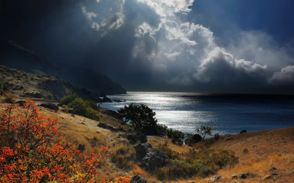 Dramatic coastline of Crimea, Russia, with dark clouds over the sea and horizon, framed by autumn foliage under a moody sky in this HD desktop wallpaper.