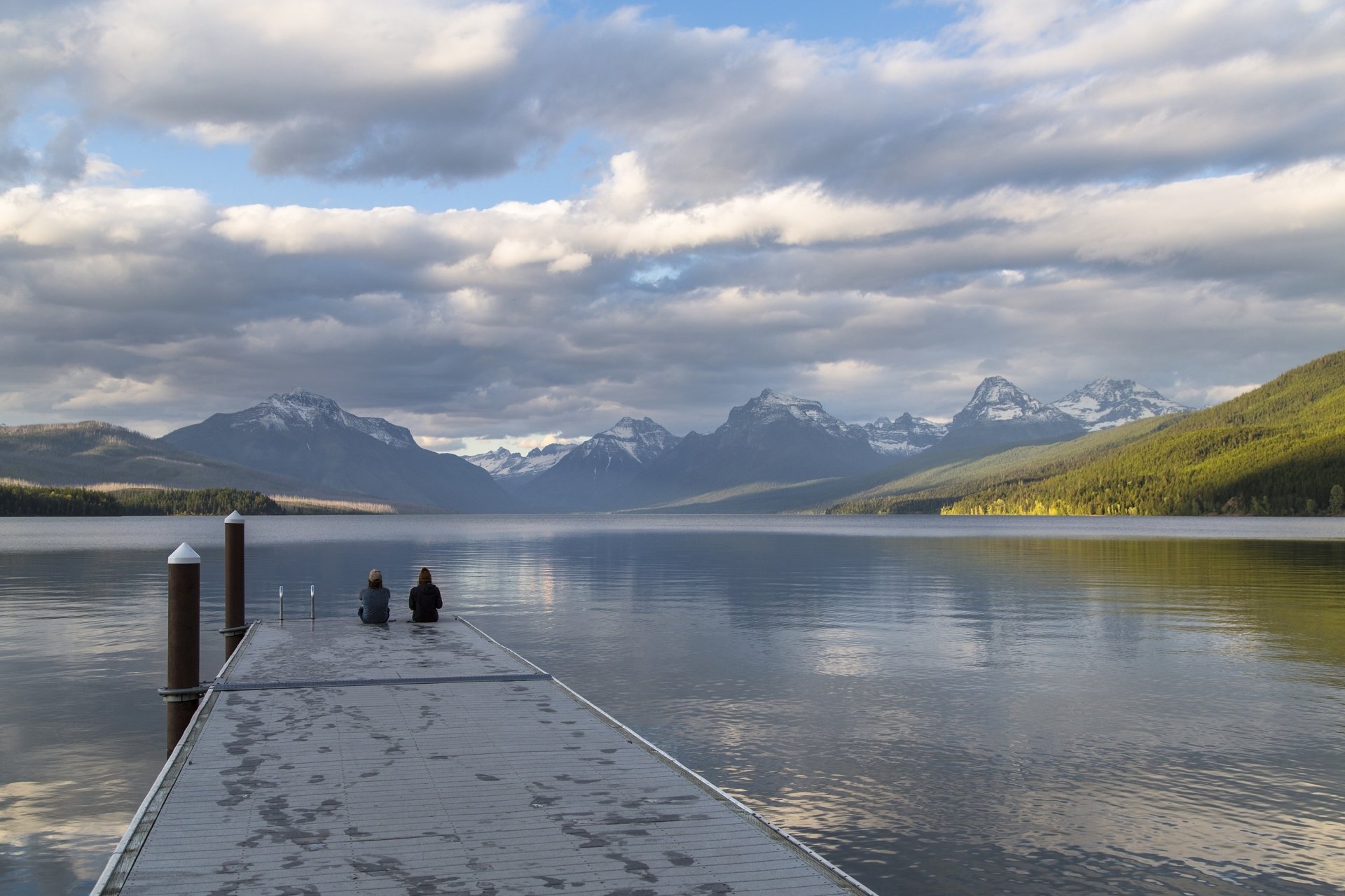 Tranquil view of Lake McDonald in Montana with a wooden pier extending into calm waters, framed by majestic mountains and a partly cloudy sky.