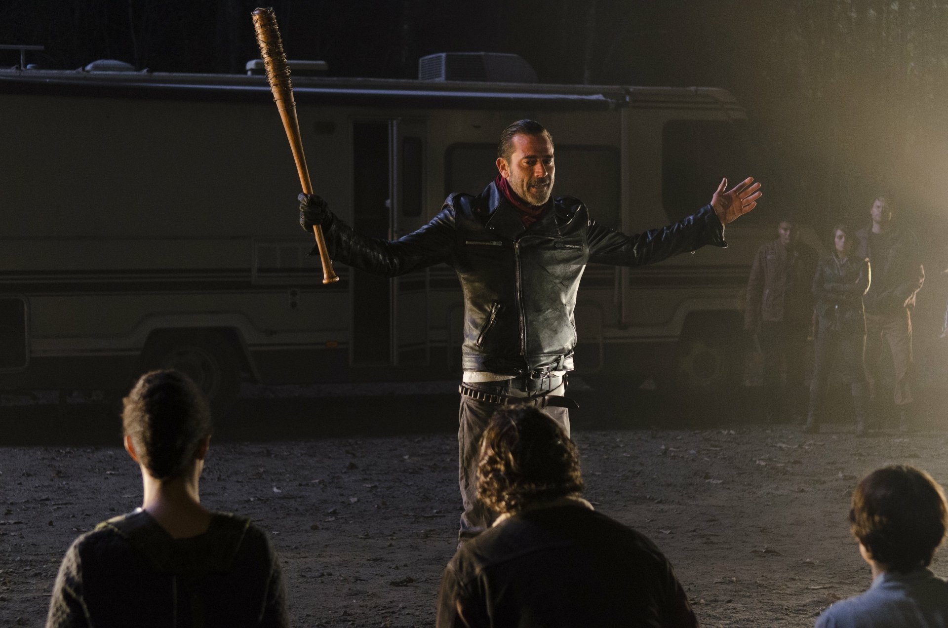 Jeffrey Dean Morgan as Negan from The Walking Dead stands confidently with a bat, addressing a group, set against a backdrop of an RV and a dark, eerie atmosphere.