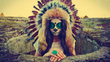 Artistic HD PC desktop wallpaper of a person with sugar skull face paint wearing a feathered headdress and turquoise ring, framed by rocky ground and a warm sunset.