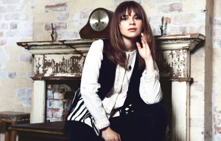 English singer Gabrielle Aplin with brunette hair and brown eyes poses indoors in front of a rustic fireplace, featured in an HD PC desktop wallpaper.