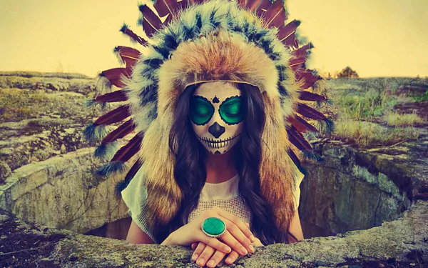 Artistic HD PC desktop wallpaper of a person with sugar skull face paint wearing a feathered headdress and turquoise ring, framed by rocky ground and a warm sunset.
