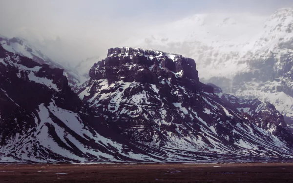 2K Quad HD PC desktop wallpaper and background: nature scene of a Snowy Mountain with rugged cliffs, streaks of snow, a cloudy sky and a barren foreground plain.