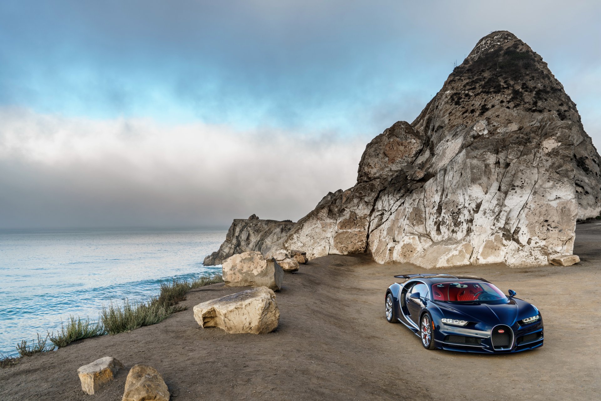 A sleek Bugatti Chiron parked along a rugged coastline, with the ocean waves lapping at the shore and dramatic cliffs in the background, creating a stunning HD desktop wallpaper.
