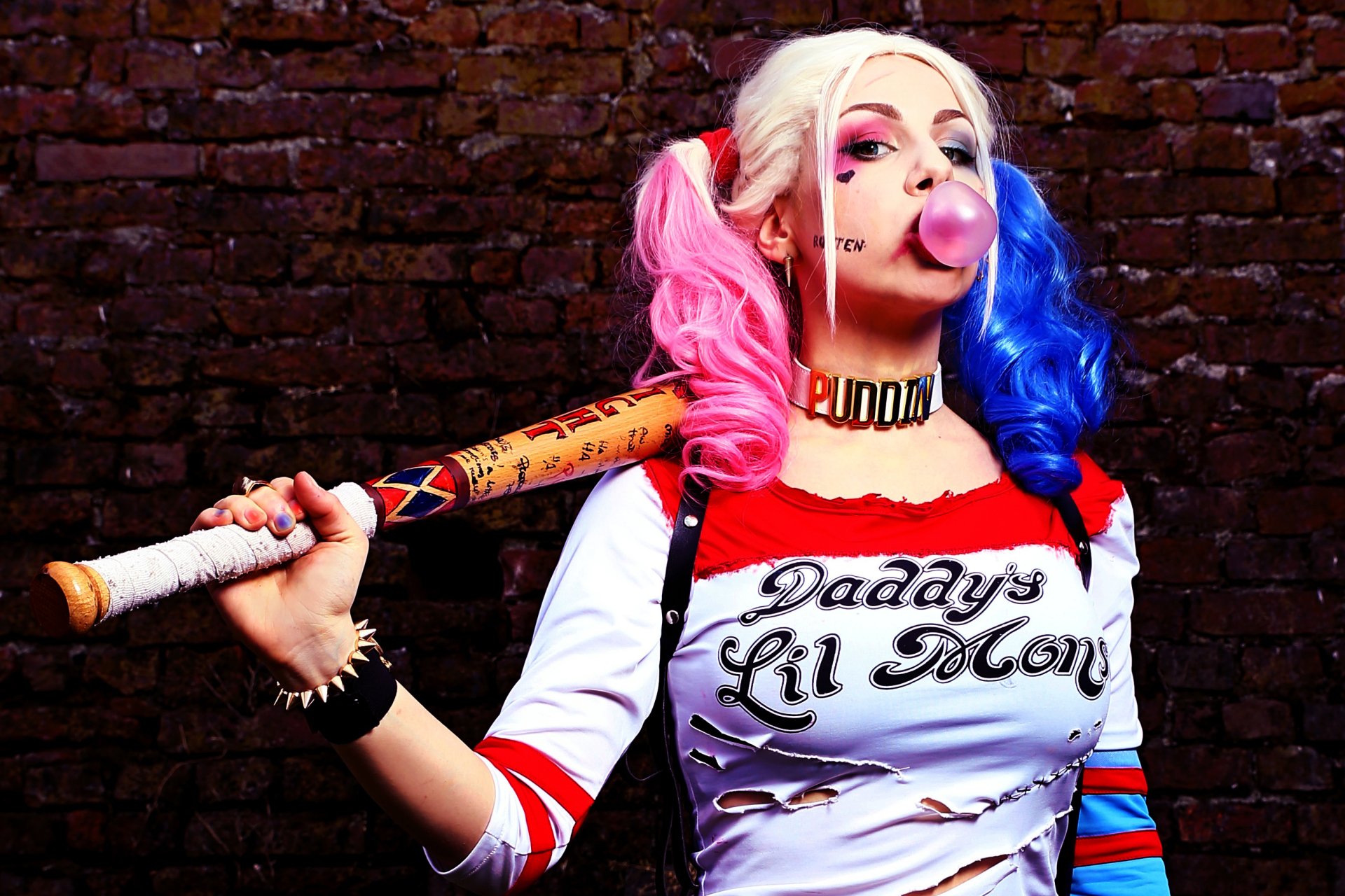 HD desktop wallpaper of a woman in Harley Quinn cosplay from Suicide Squad, blowing a bubble, holding a baseball bat, with pink and blue pigtails, against a brick wall background.