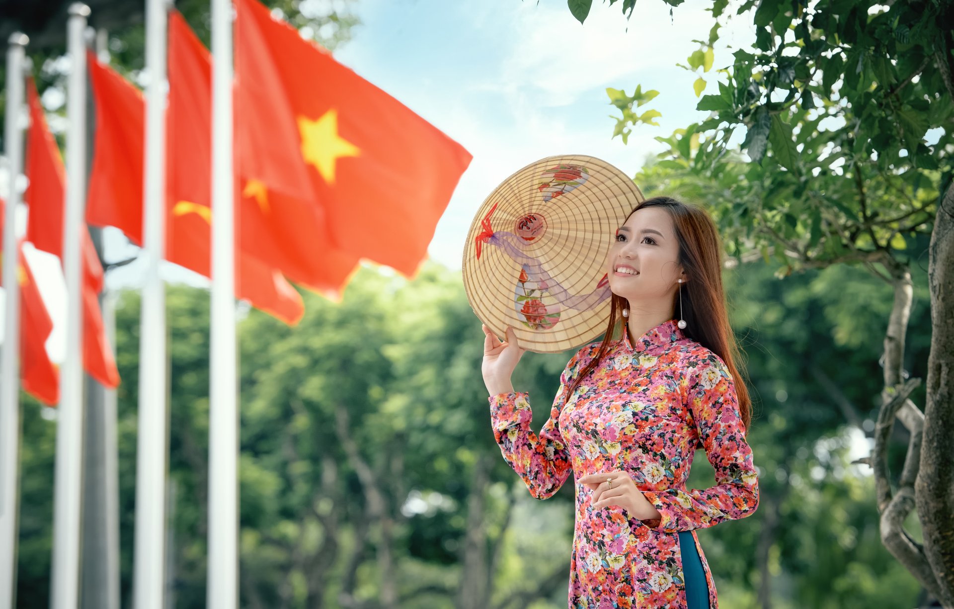 A smiling woman with brown eyes and brunette hair wears a traditional costume and hat, standing among flags, showcasing cultural beauty in a vibrant outdoor setting.