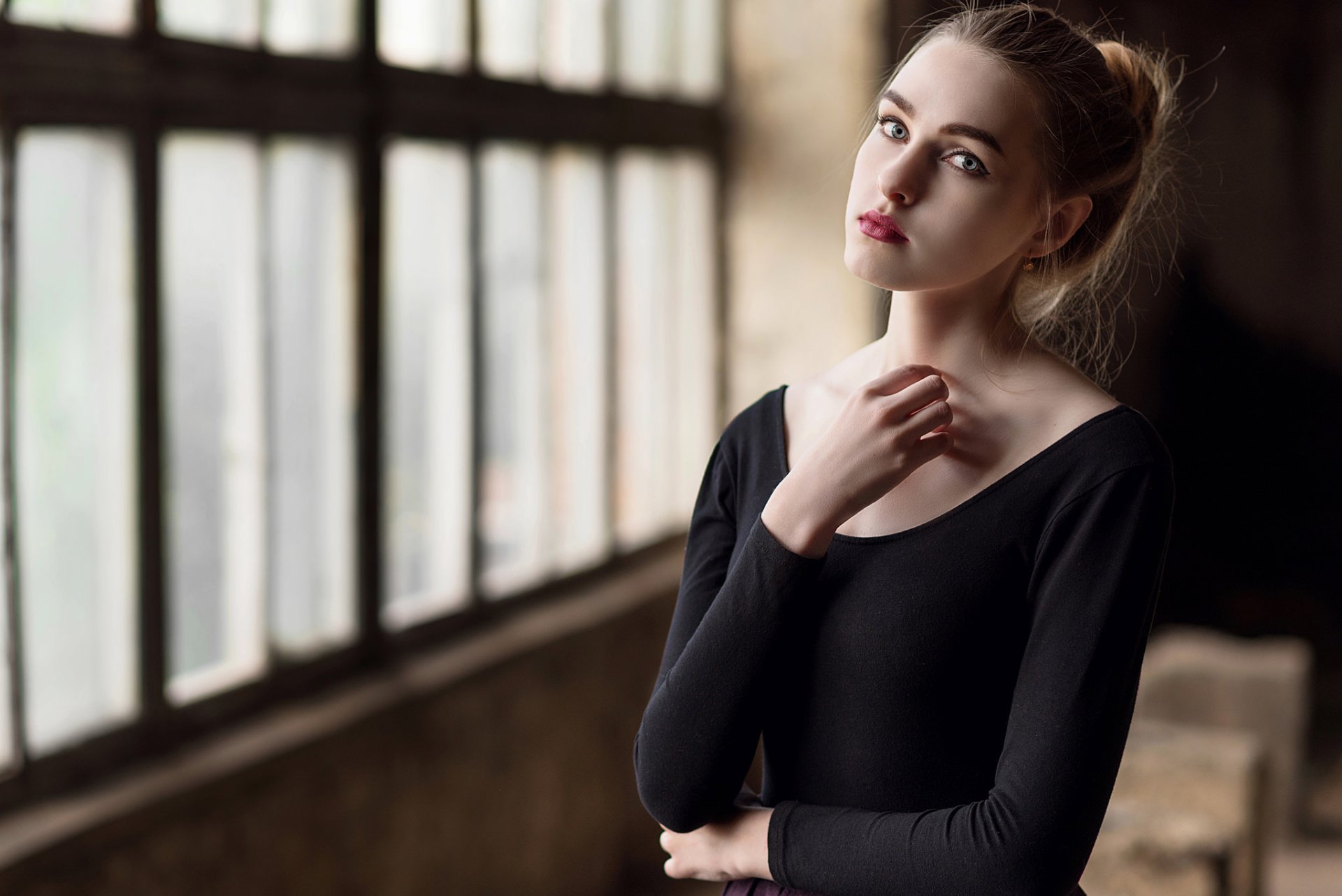 HD PC desktop wallpaper: lipstick-wearing woman model with blue eyes in a black top, gazing by a large industrial window.