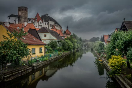 A serene canal in a picturesque Austrian village, featuring colorful houses and lush greenery, all reflected in the calm water under a dramatic, cloudy sky. Perfect HD wallpaper.