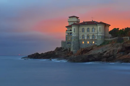 HD wallpaper of a historic lighthouse building on the rocky coast of Livorno, Italy, overlooking the calm ocean at sunset with a colorful sky.